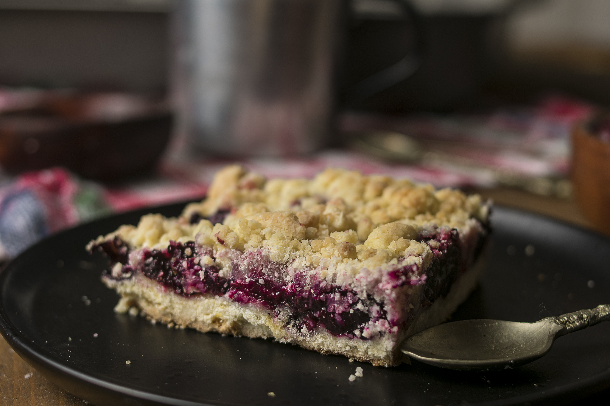 Slice of Italian berry crumble tart on a black plate, with a bowl of frozen mixed berries and the remaining bake softly blurred in the background.